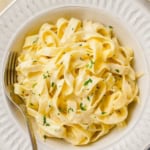 A bowl of creamy fettuccine Alfredo garnished with chopped parsley, served with a fork on a white plate.