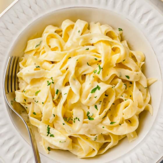 A bowl of creamy fettuccine Alfredo garnished with chopped parsley, served with a fork on a white plate.