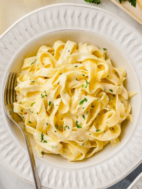 A bowl of creamy fettuccine Alfredo garnished with chopped parsley, served with a fork on a white plate.