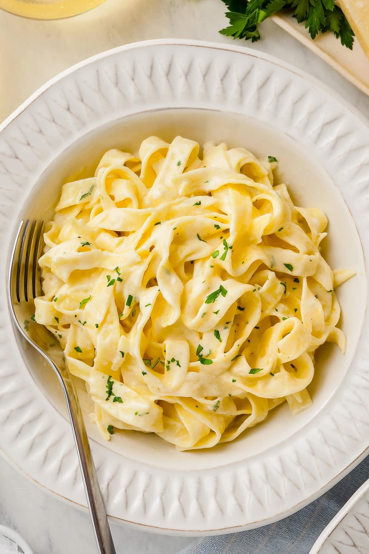 A bowl of creamy fettuccine Alfredo garnished with chopped parsley, served with a fork on a white plate.