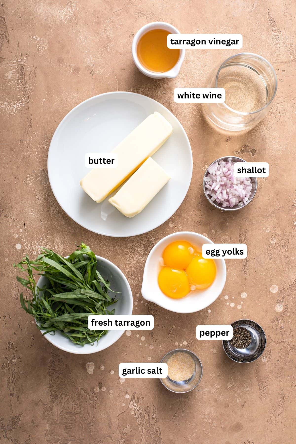 This easy Béarnaise Sauce recipe is made in your food processor, or blender, for an easy foolproof method! It’s creamy, rich and perfect! Overhead view of labeled ingredients for easy béarnaise sauce recipe: two butter sticks on a plate, chopped shallot, tarragon vinegar, white wine, three egg yolks, fresh tarragon, garlic salt, and pepper on a tan surface.