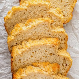 Six slices of golden, rustic beer bread are arranged on white parchment paper. The bread has a fluffy, slightly crumbly interior and a crisp, lightly browned crust.