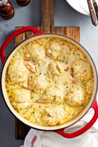 A red Dutch oven filled with creamy homemade chicken and dumplings, featuring fluffy dumplings floating in a thick, herb-speckled broth with visible pieces of carrots and chicken.