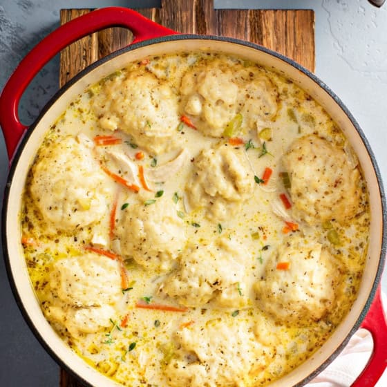 A red Dutch oven filled with creamy homemade chicken and dumplings, featuring fluffy dumplings floating in a thick, herb-speckled broth with visible pieces of carrots and chicken.