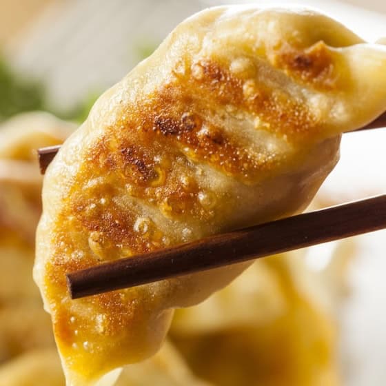 A pair of chopsticks holds a golden-brown, pan-fried Chinese dumpling above a plate filled with more dumplings in the background.