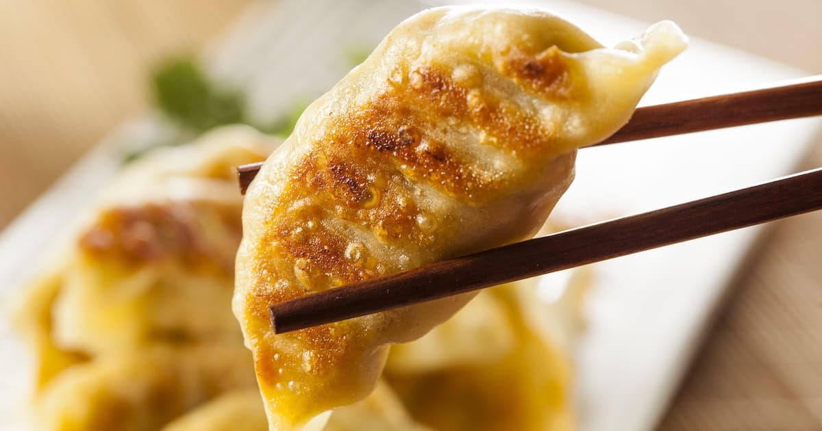 A pair of chopsticks holds a golden-brown, pan-fried Chinese dumpling above a plate filled with more dumplings in the background.