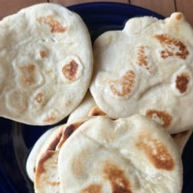 A close-up of several pieces of homemade pita bread with golden brown spots, arranged on a dark blue plate. The bread appears soft and slightly puffy.