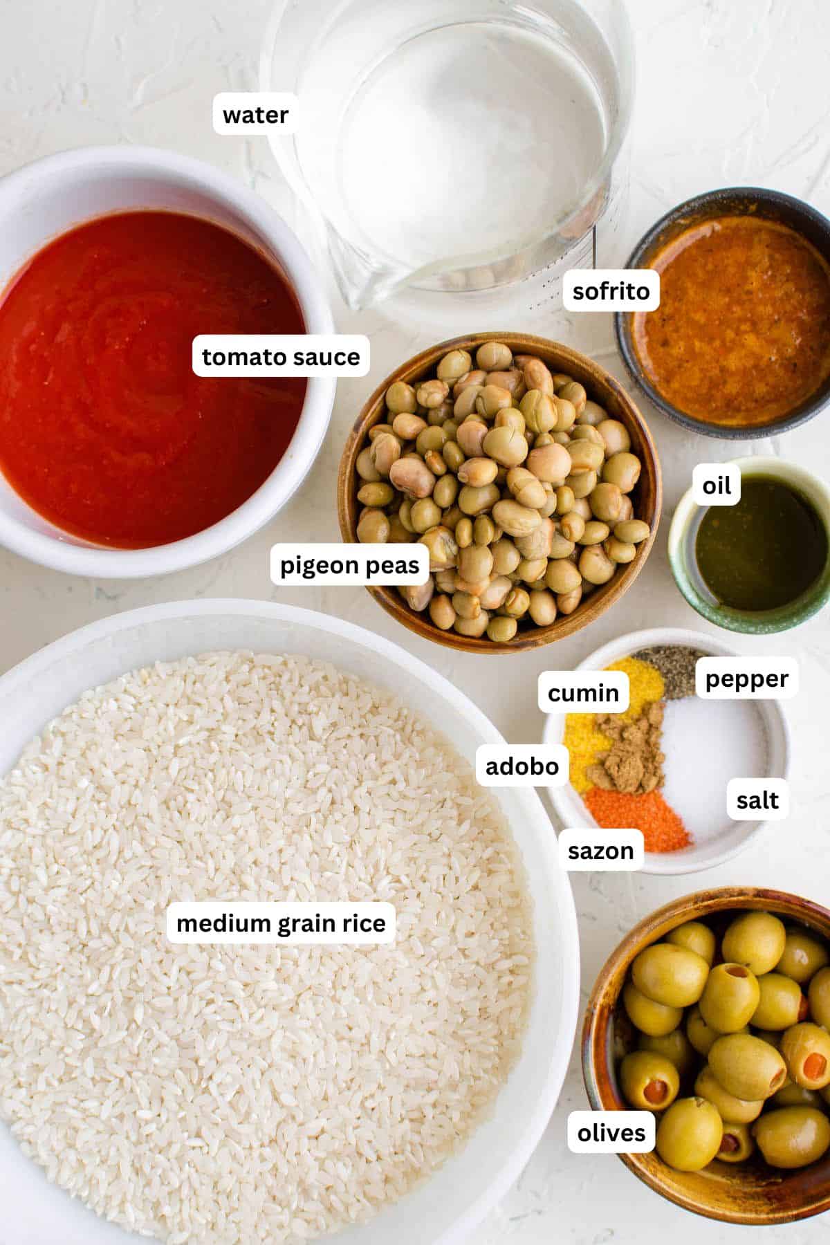 Bowls and cups containing labeled ingredients for arroz con gandules recipe: water, tomato sauce, sofrito, oil, pigeon peas, cumin, pepper, salt, adobo, sazon, medium grain rice, and olives on a white surface.
