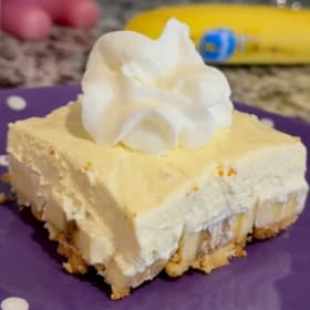 A slice of banana pudding bars with a Nilla wafter crust, layered banana slices, creamy banana pudding filling, and a swirl of whipped cream on top, served on a purple plate with white polka dots.