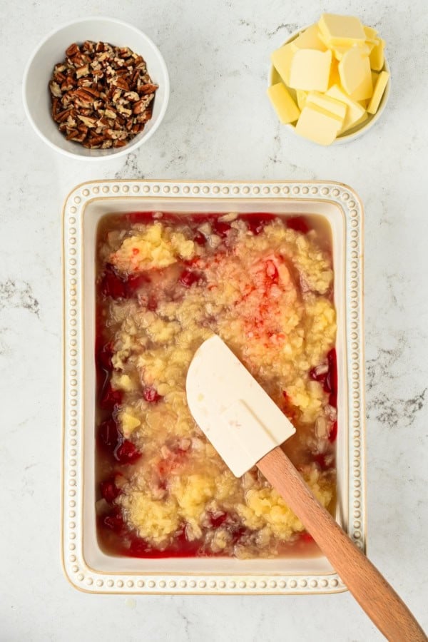 A baking dish with an unbaked mixture of crushed pineapple and strawberry pie filling being stirred with a spatula. Nearby are bowls of chopped pecans and sliced butter on a white countertop.