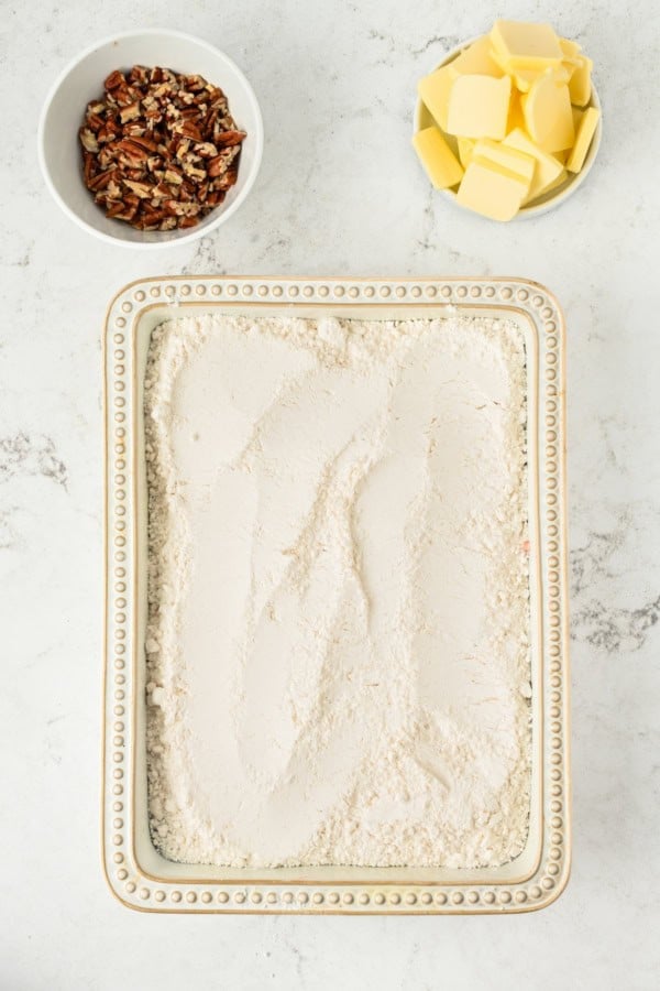 A rectangular baking dish filled with cake mix sits on a white surface. Above it, there is a bowl of chopped pecans on the left and a bowl of cubed butter on the right.