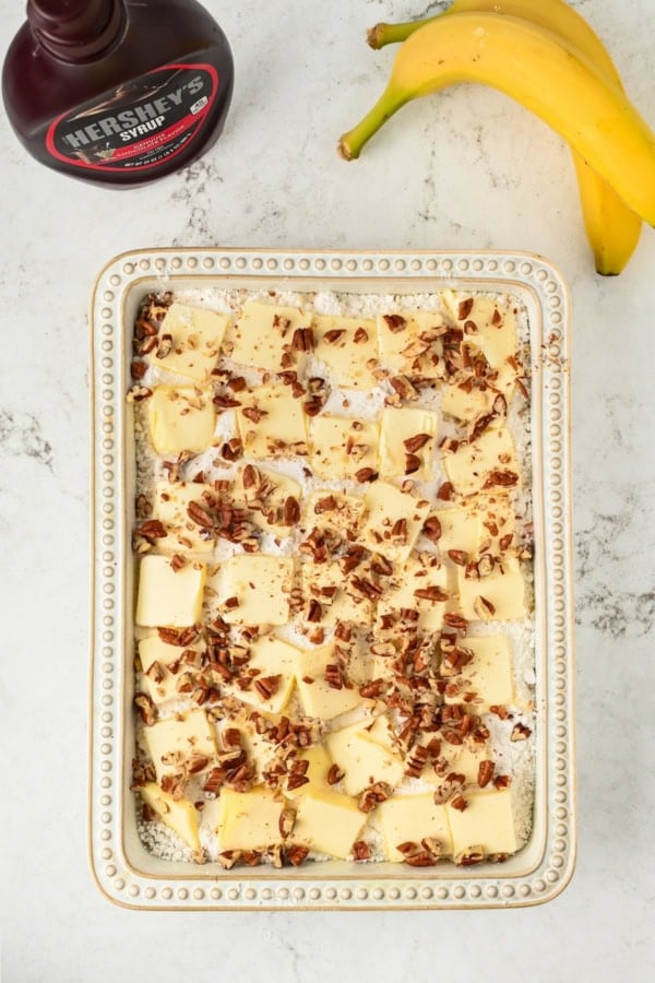 Rectangular baking dish filled with cubed butter, chopped pecans, and other ingredients. A bottle of Hershey’s syrup and two bananas are nearby on a marble countertop.