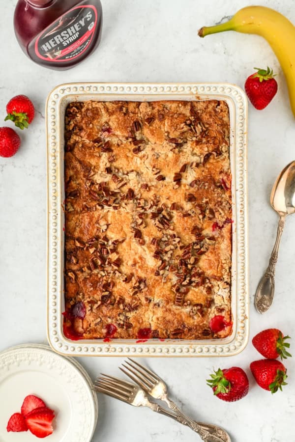 A baked banana split cake topped with chopped pecans in a rectangular dish, surrounded by strawberries, a banana, a bottle of Hershey’s syrup, plates, forks, and a spoon on a white marble surface.