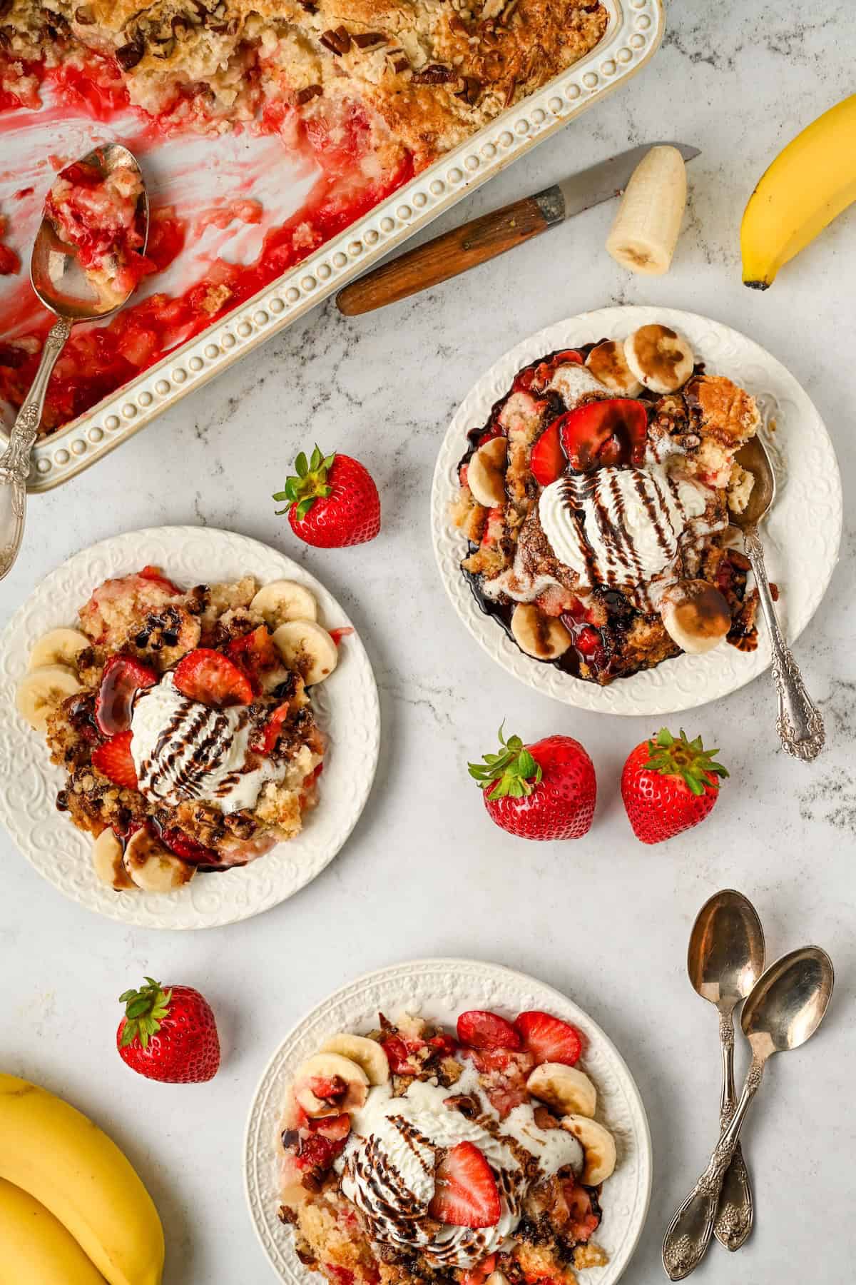 Three plates of banana split dump cake topped with whipped cream, chocolate drizzle, sliced bananas, and strawberries are arranged on a white surface, surrounded by fresh strawberries, bananas, and a partially served baking dish.