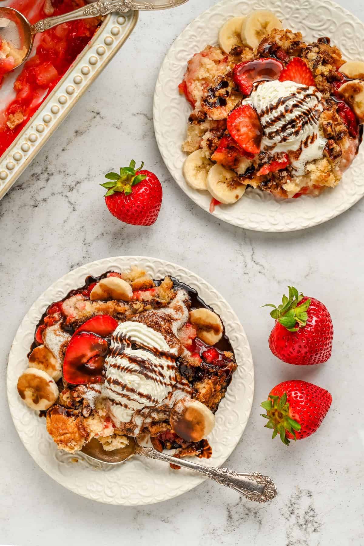 Two plates of banana split cake topped with sliced bananas, strawberries, vanilla ice cream, and chocolate sauce sit on a white surface, surrounded by fresh strawberries and a baking dish of the same dessert.