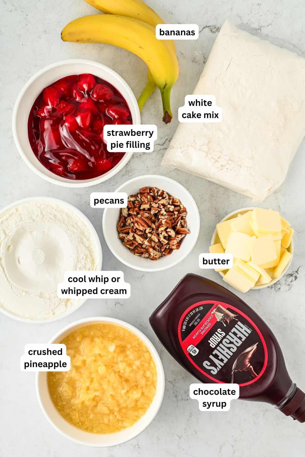 Labeled ingredients for making banana split dump cake recipe on a countertop: bananas, white cake mix, strawberry pie filling, cool whip, crushed pineapple, chopped pecans, butter slices, and a bottle of chocolate syrup.