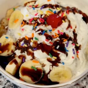 A bowl of banana split cake topped with whipped cream, chocolate syrup, rainbow sprinkles, banana slices, and a cherry.