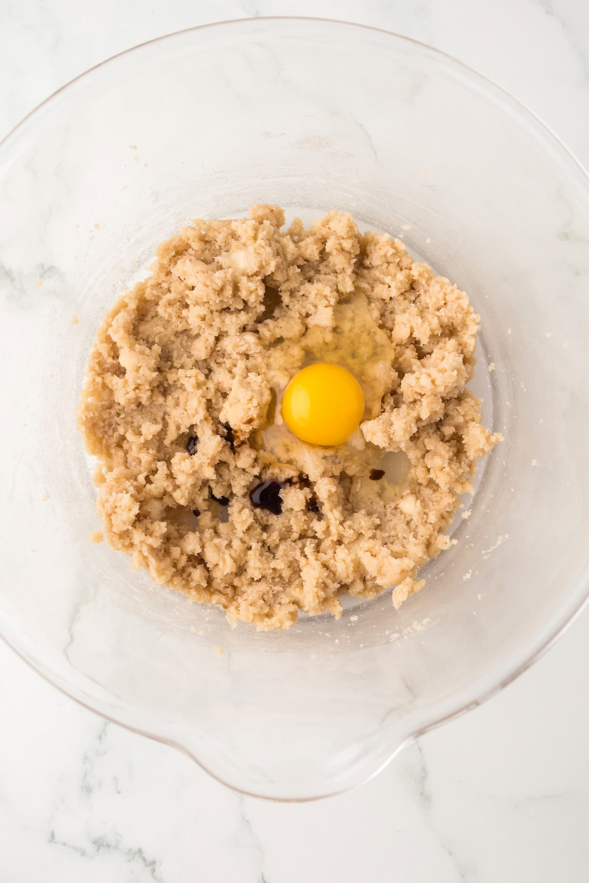 A clear glass mixing bowl with cookie dough mixture and a whole raw egg on top, sitting on a white marble surface.