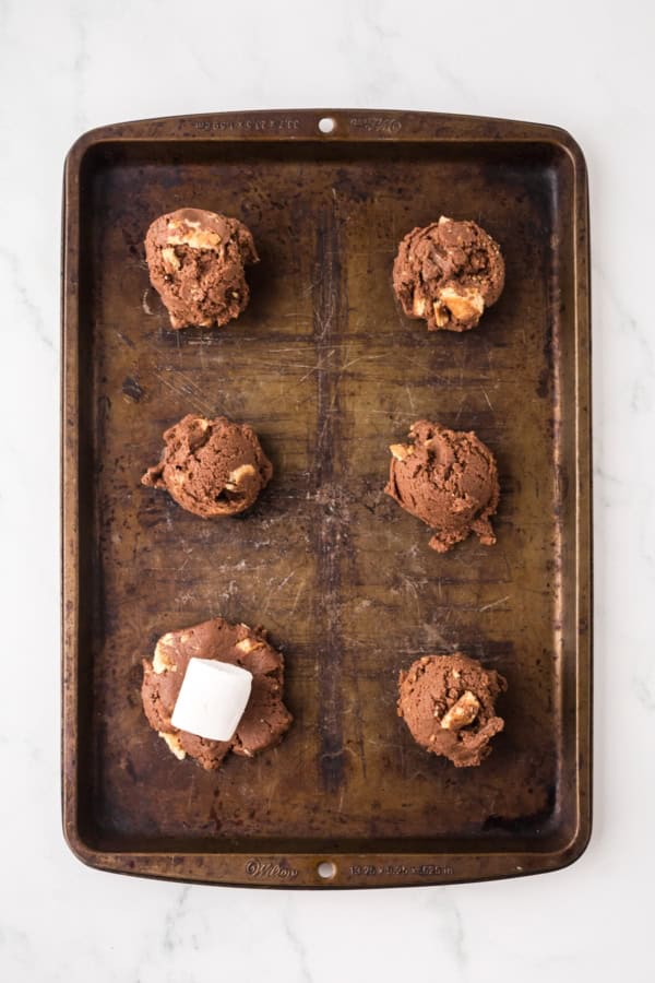 Six scoops of chocolate cookie dough, one topped with a marshmallow, are spaced evenly on a worn metal baking sheet set on a white marble surface.