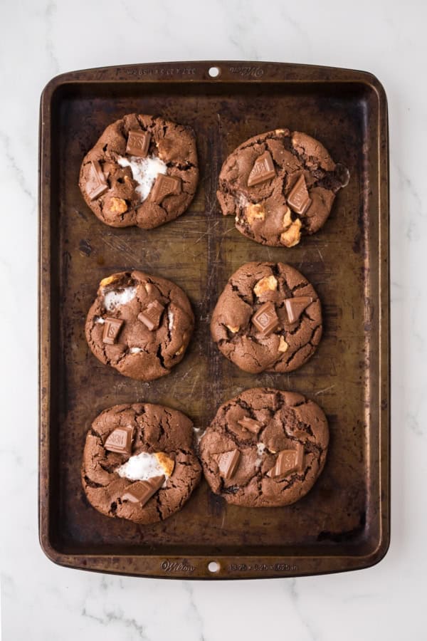 A baking tray with six large chocolate s'mores cookies, some with visible chunks of marshmallow and pieces of chocolate on top, placed on a light marble surface.