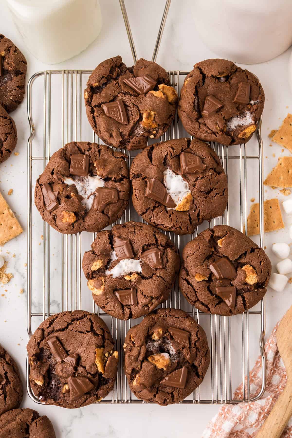 A cooling rack with nine chocolate marshmallow cookies topped with chunks of chocolate, melted marshmallow, and bits of graham cracker. Scattered marshmallows, graham crackers, and a glass of milk are nearby.