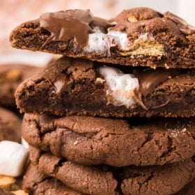 A stack of chocolate s'mores cookies with gooey marshmallow and melted chocolate inside. One cookie on top is broken in half, showing the soft, melty filling. Marshmallow pieces and crumbs are visible nearby.
