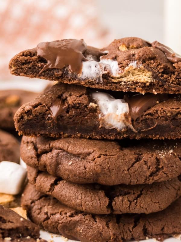 A stack of chocolate s'mores cookies with gooey marshmallow and melted chocolate inside. One cookie on top is broken in half, showing the soft, melty filling. Marshmallow pieces and crumbs are visible nearby.
