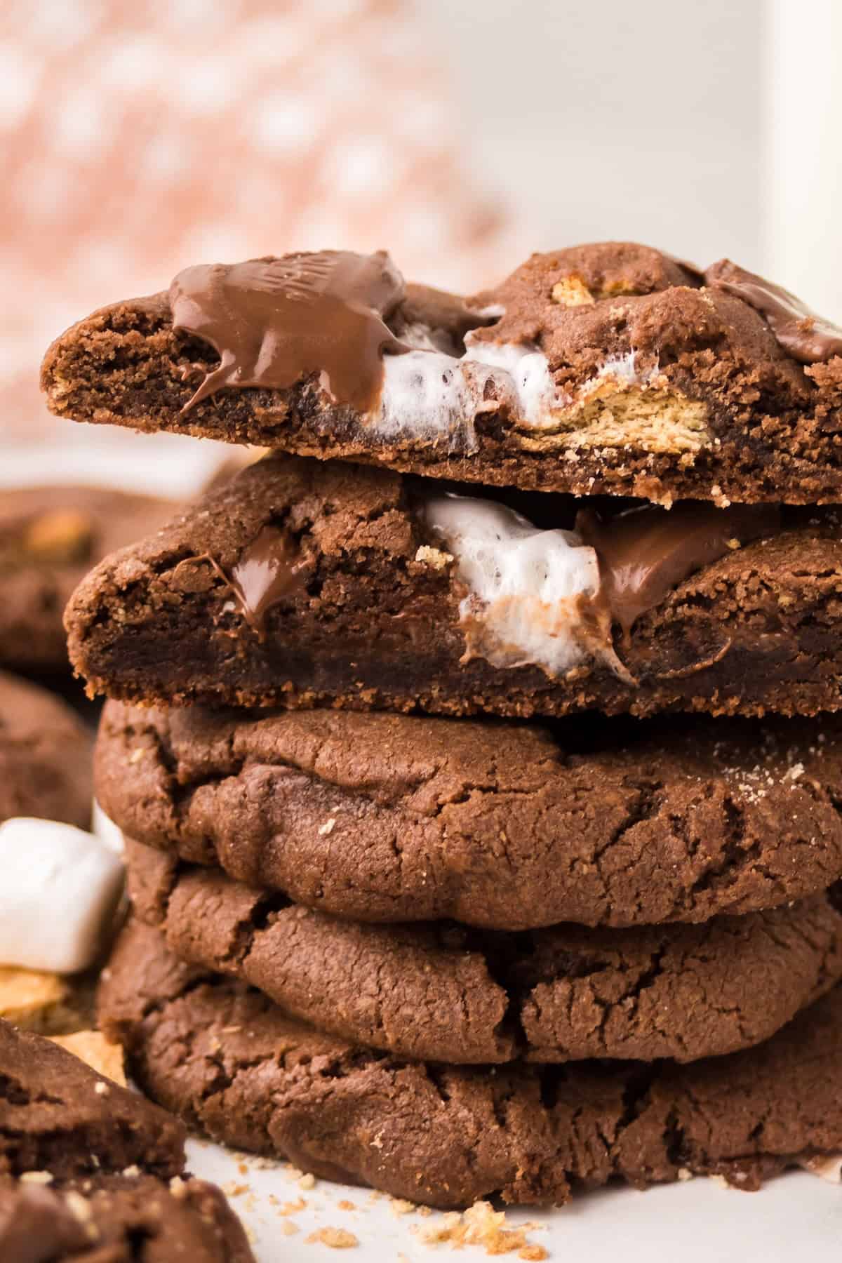 A stack of chocolate s'mores cookies with gooey marshmallow and melted chocolate inside. One cookie on top is broken in half, showing the soft, melty filling. Marshmallow pieces and crumbs are visible nearby.