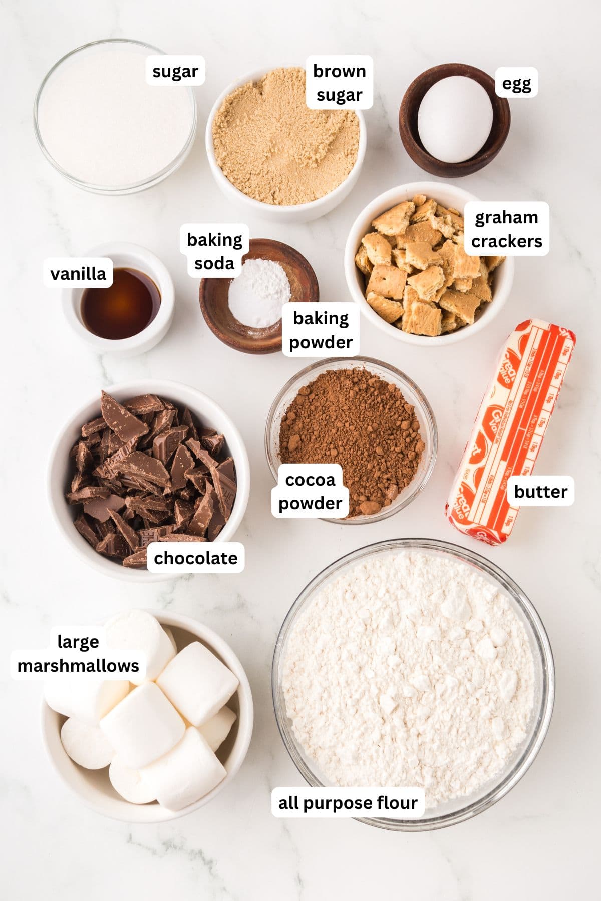 Ingredients for baking chocolate s'mores cookies are arranged on a white surface, each labeled: sugar, brown sugar, egg, baking soda, baking powder, graham crackers, vanilla, chocolate, cocoa powder, butter, all purpose flour, and large marshmallows.