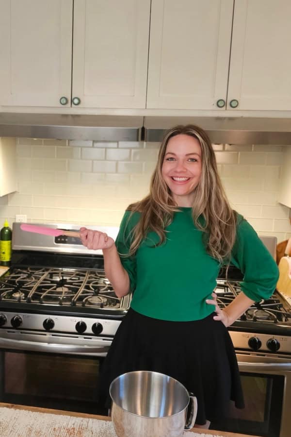 A smiling woman in a green top stands in a kitchen, holding a pink spatula next to a metal mixing bowl on a counter, with a stove and white cabinets in the background.
