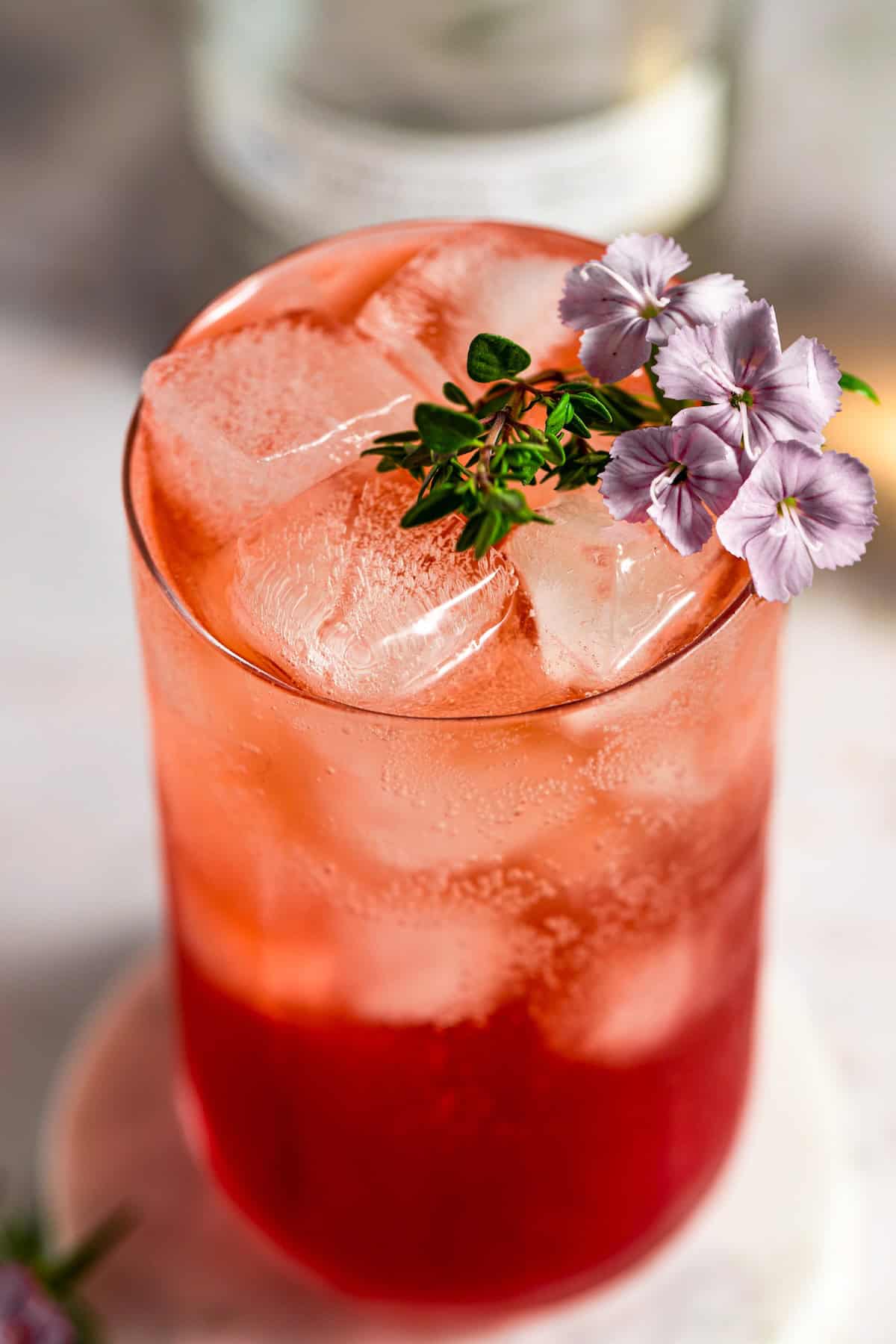 A vibrant red iced transfusion drink in a glass, garnished with small purple flowers and a sprig of greenery, sits on a light surface. Large ice cubes fill the glass, and the background is softly blurred.