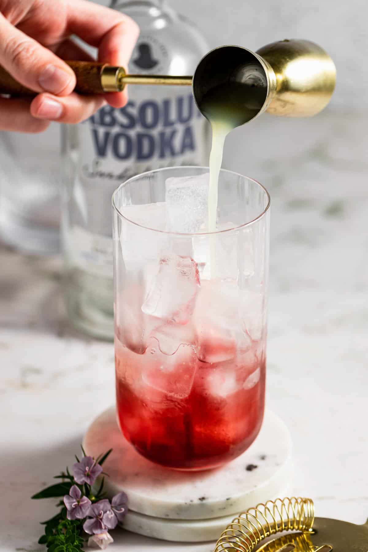 A hand pours lime juice from a jigger into a glass filled with ice and grape juice, with a bottle of vodka in the background and small purple flowers and a gold strainer nearby.