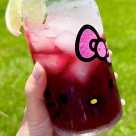 A hand holds a hello kitty glass filled with a transfusion drink (a purple iced drink) and garnished with a slice of lime. The background shows bright green grass in sunlight.