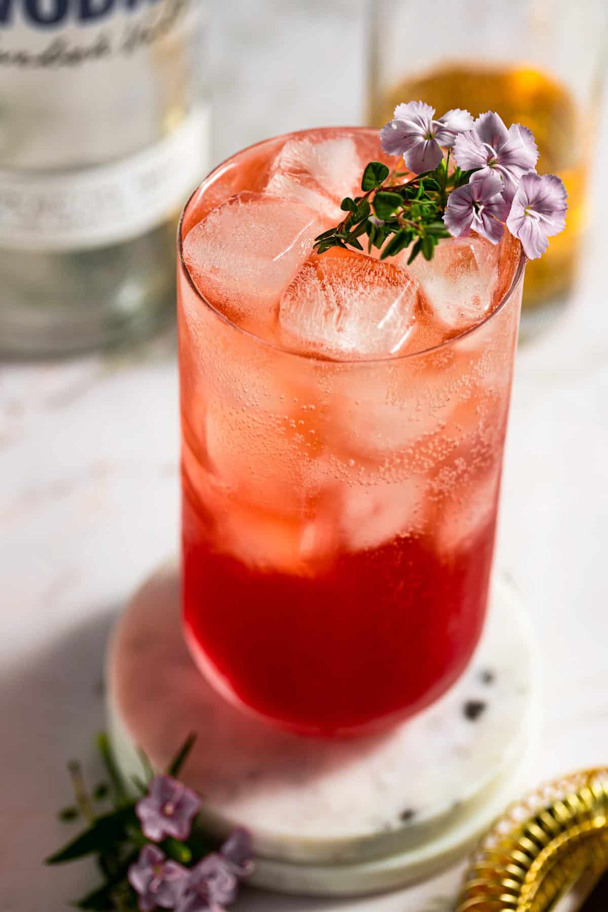 A tall glass filled with a transfusion cocktail, garnished with small purple flowers and herbs, sits on a round white marble coaster.