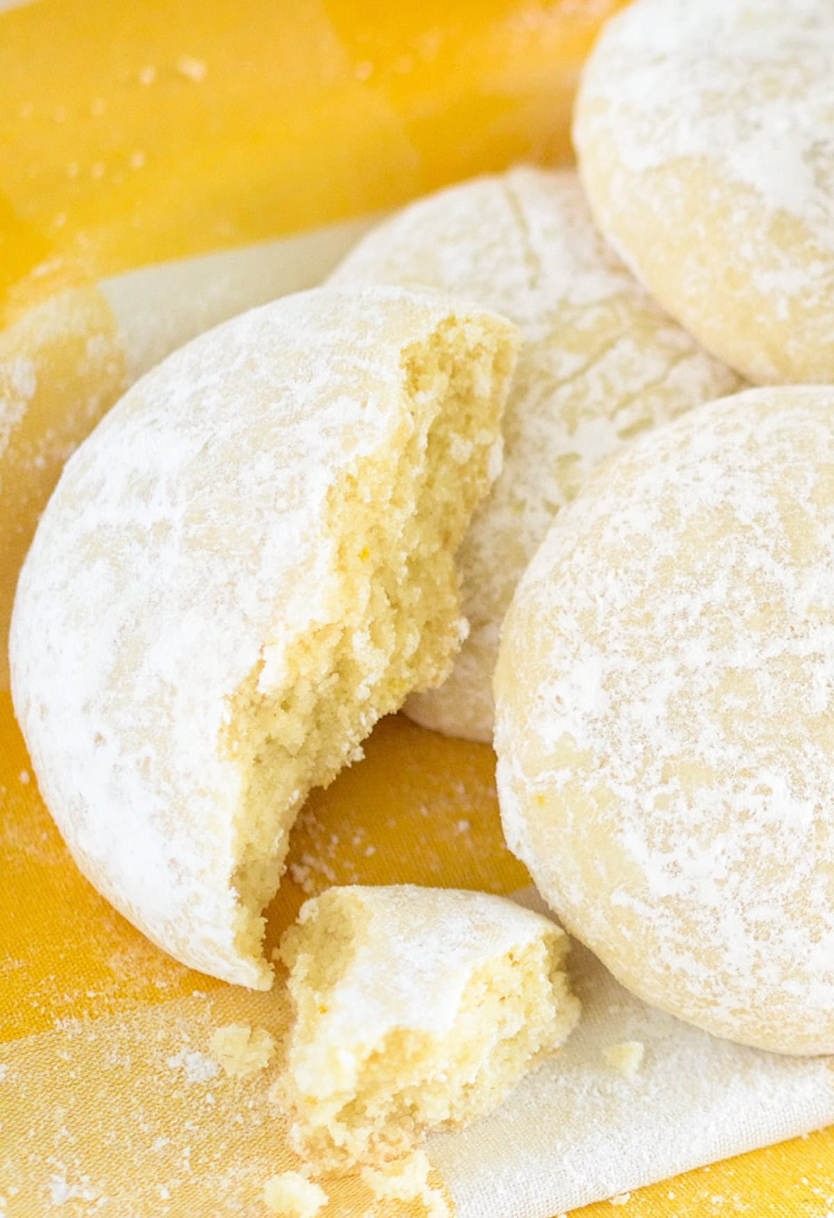Four round, light yellow lemon cooler cookies dusted with powdered sugar are shown. One cookie has a bite taken out, with a piece and scattered crumbs in front of it, all on a yellow and white background.