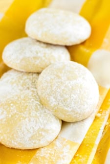 Four round, light yellow lemon cooler cookies dusted with powdered sugar are arranged on a yellow and white striped cloth.