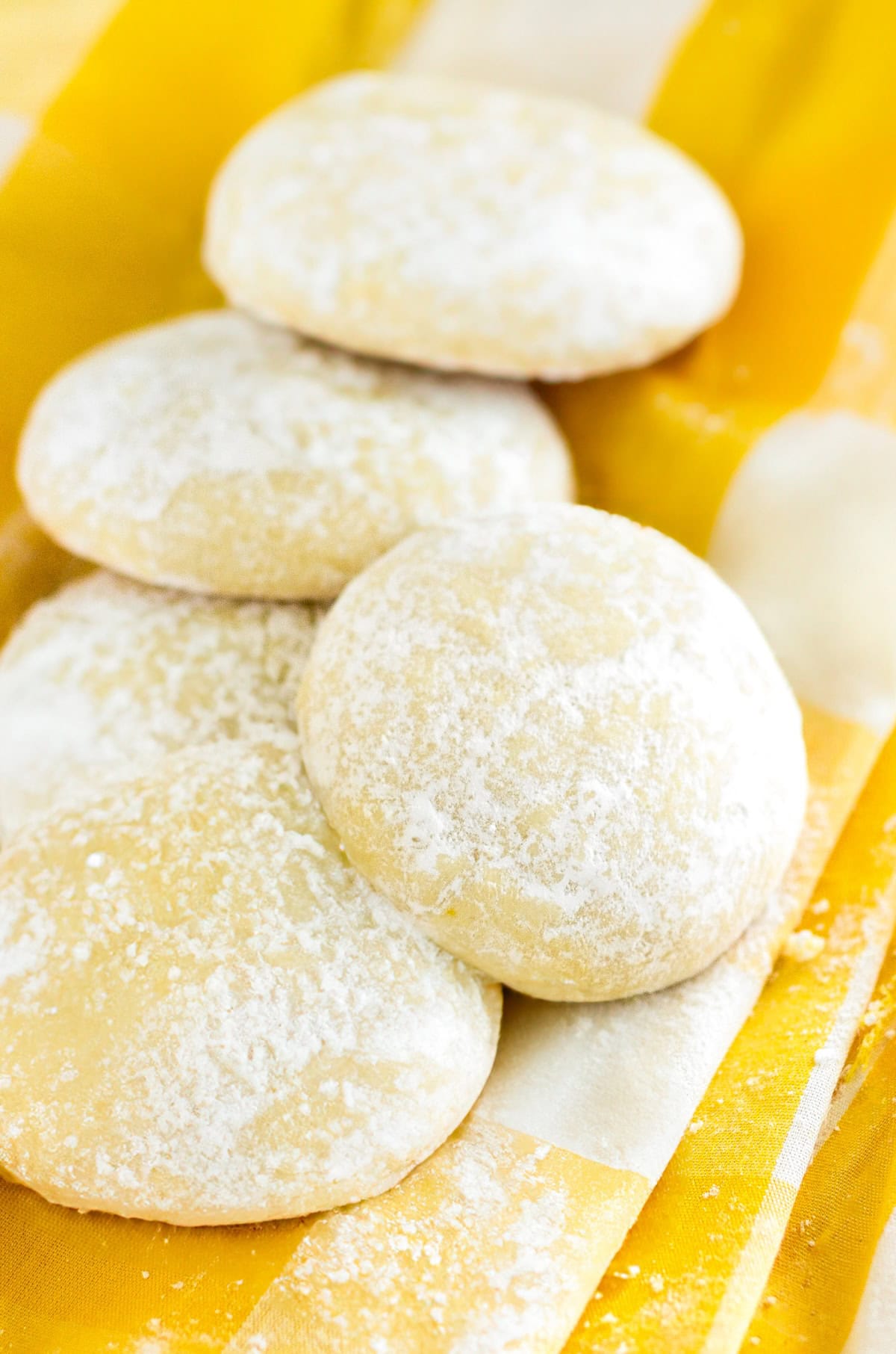 Four round, light yellow lemon cooler cookies dusted with powdered sugar are arranged on a yellow and white striped cloth.