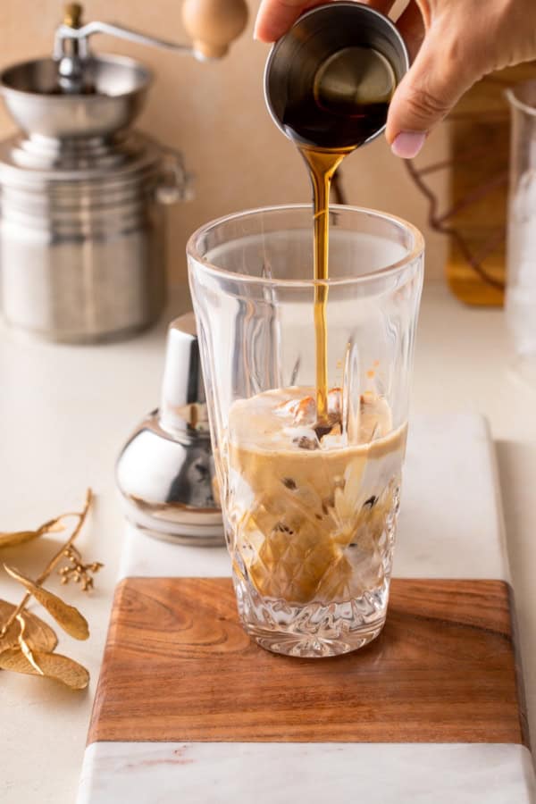 A hand pours maple syrup from a small metal cup into a tall glass filled with ice, espresso, pumpkin, and milk on a wooden board, with a metal shaker and kitchen items in the background.