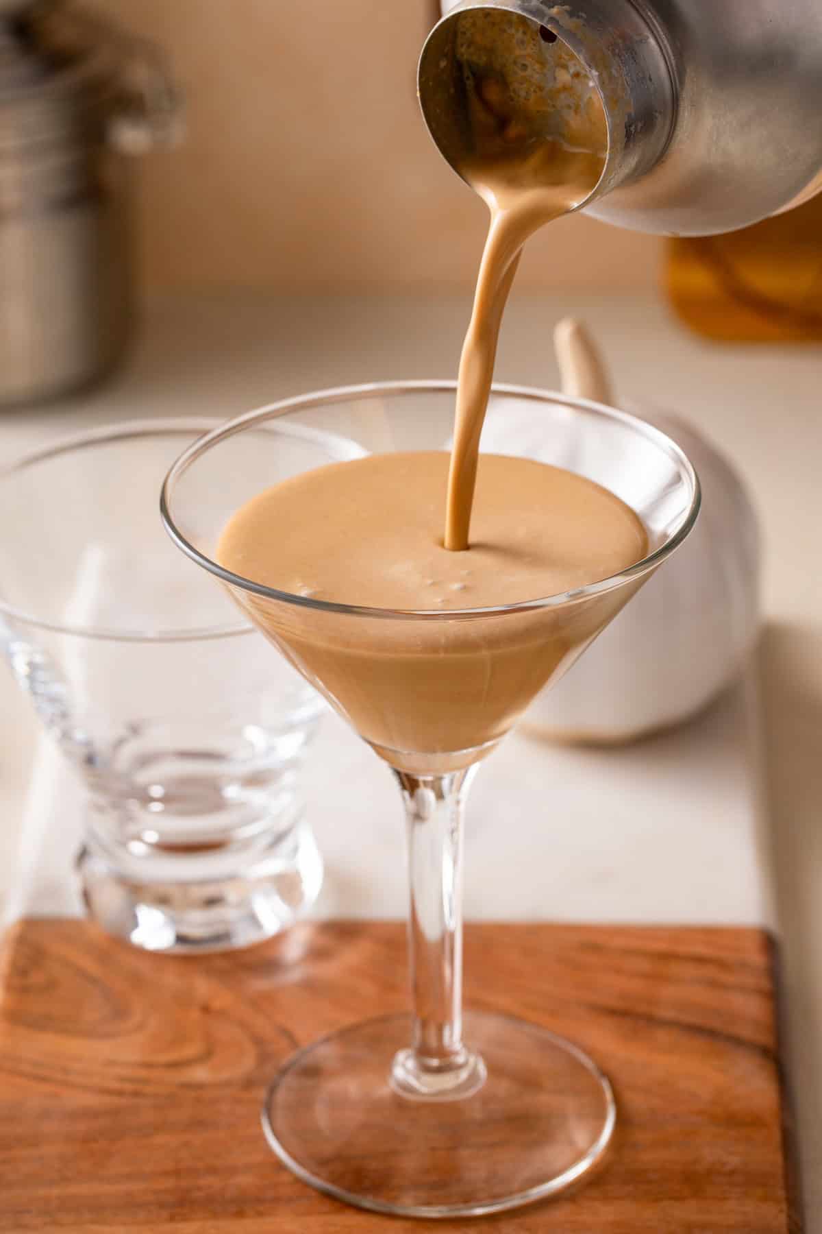 A creamy pumpkin espresso martini is being poured from a metal shaker into a martini glass on a wooden board, with an empty glass and kitchen items in the background.