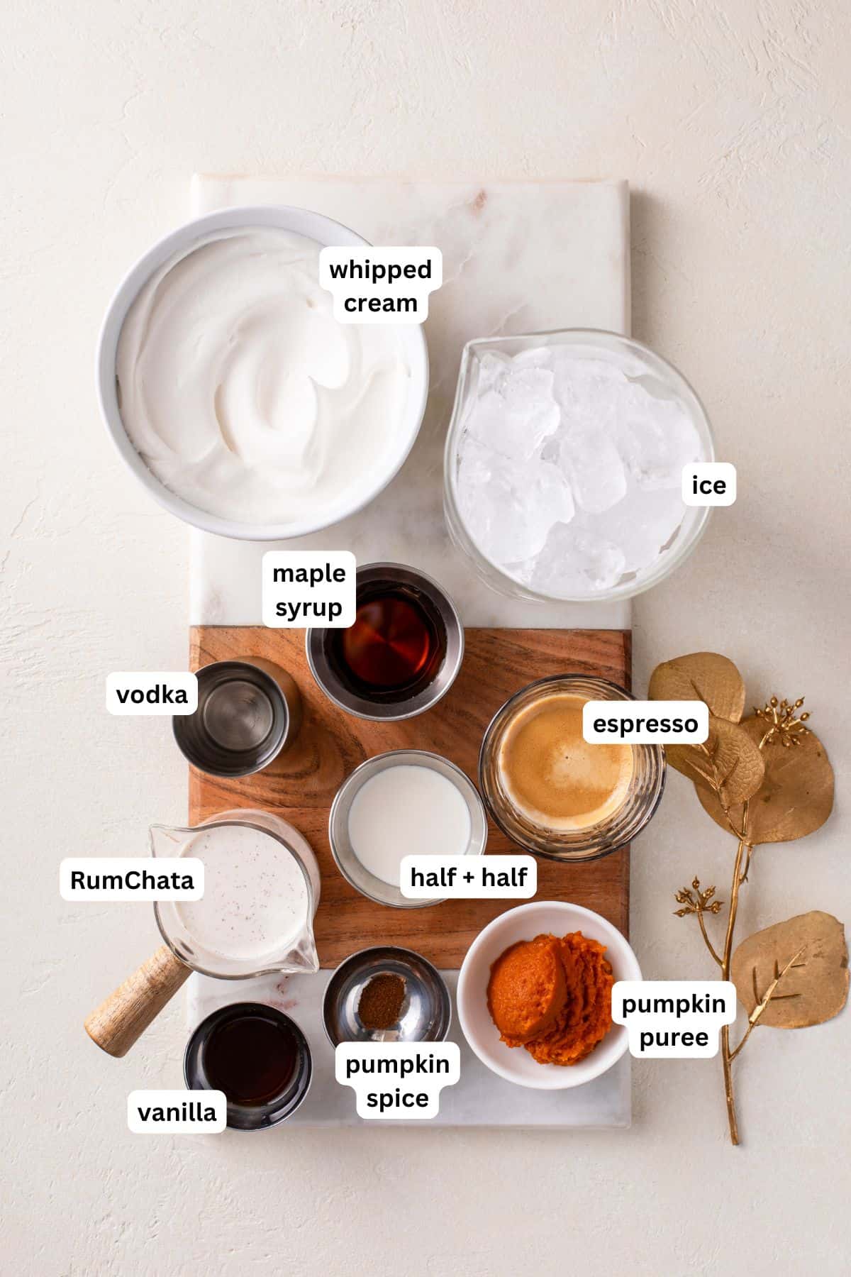 A flat lay of ingredients labeled for a pumpkin martini recipe: whipped cream, ice, vodka, RumChata, maple syrup, half & half, espresso, pumpkin puree, pumpkin spice, and vanilla, arranged on a marble and wood board.