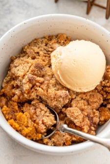 A bowl of sweet potato cobbler with a cinnamon crumb topping with a scoop of vanilla ice cream, with a spoon resting inside the bowl.
