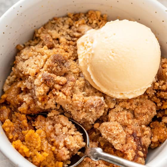 A bowl of sweet potato cobbler with a cinnamon crumb topping with a scoop of vanilla ice cream, with a spoon resting inside the bowl.