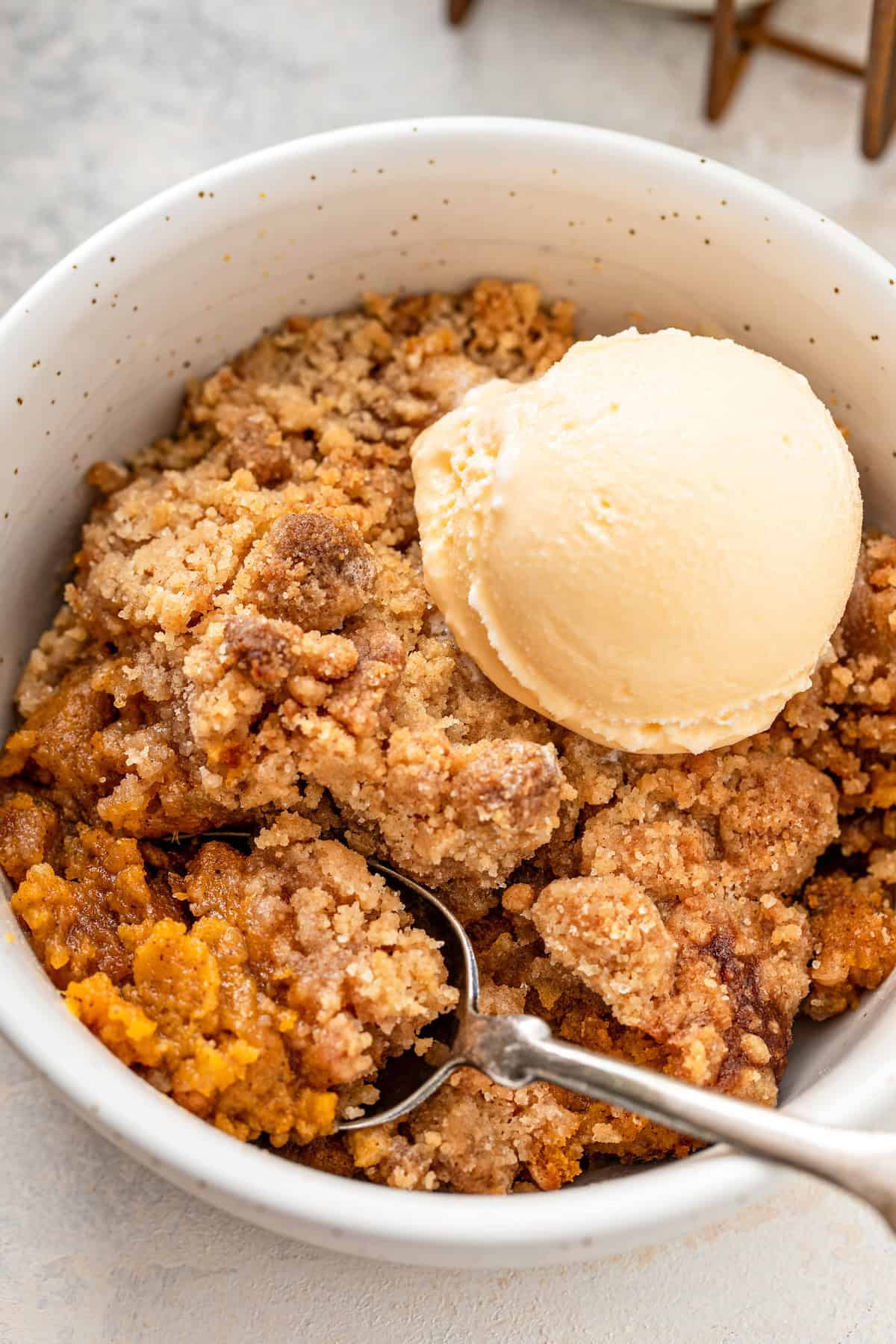 A bowl of sweet potato cobbler with a cinnamon crumb topping with a scoop of vanilla ice cream, with a spoon resting inside the bowl.