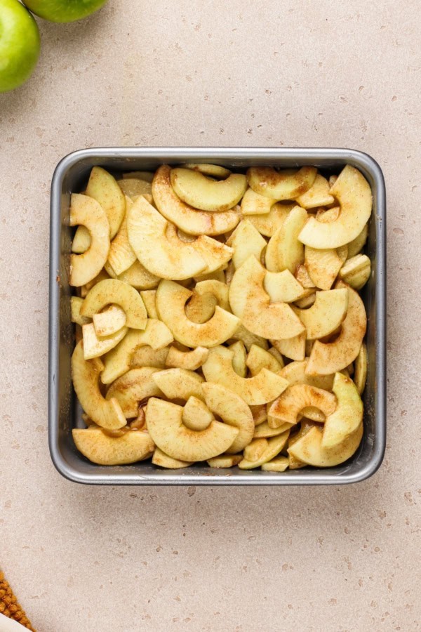 A metal square baking pan filled with sliced apples coated in cinnamon and sugar sits on a light-colored countertop. Green apples are partially visible in the top left corner.