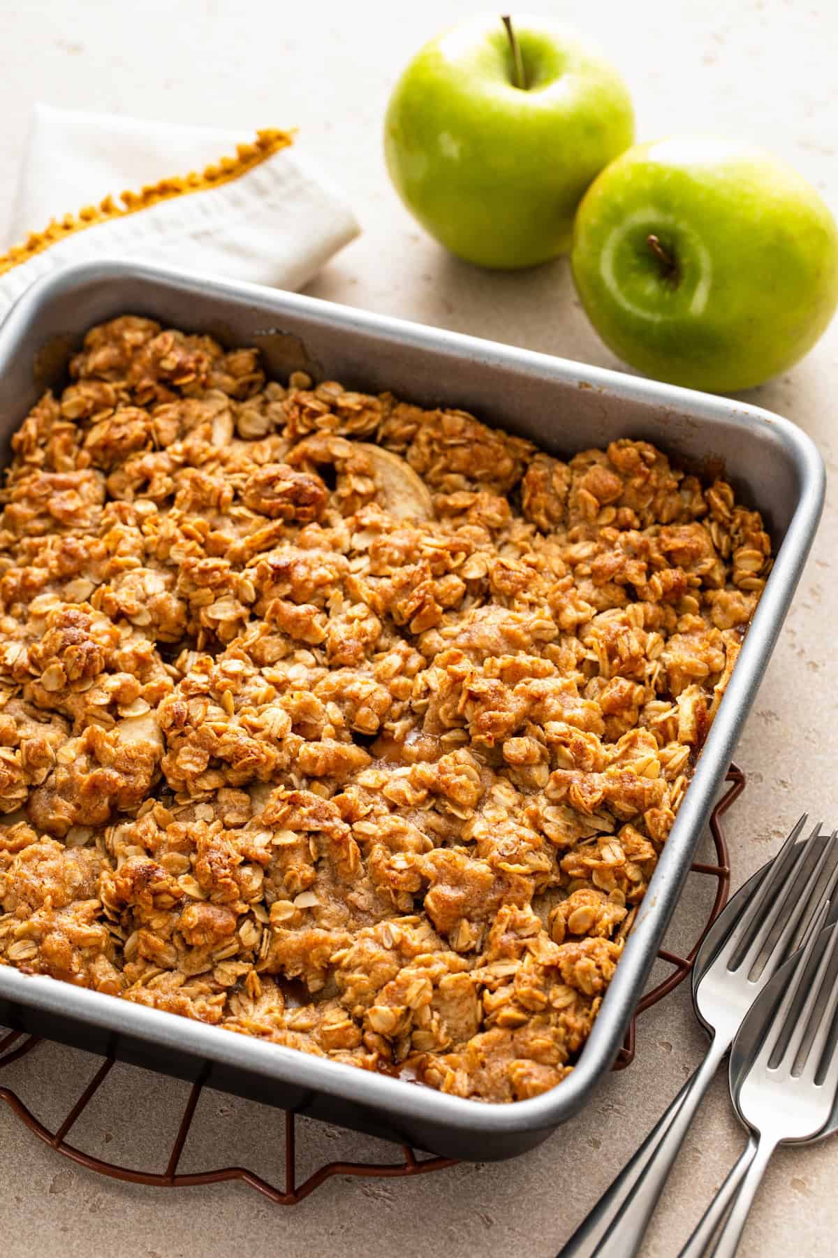 A baked apple crisp in a square metal pan sits on a cooling rack, with a golden oat topping.
