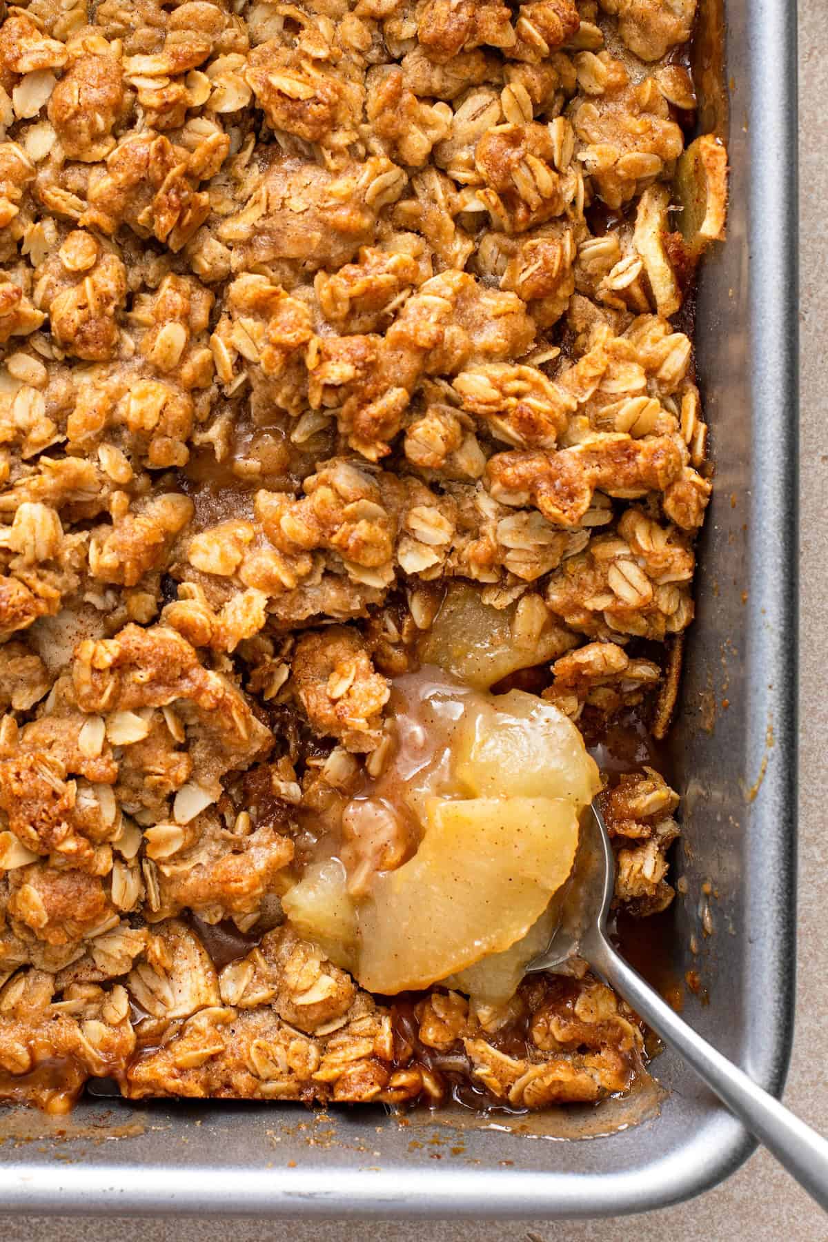 A close-up of a baked apple crisp in a metal pan, showing a golden oat topping and a spoon scooping out tender apple filling.