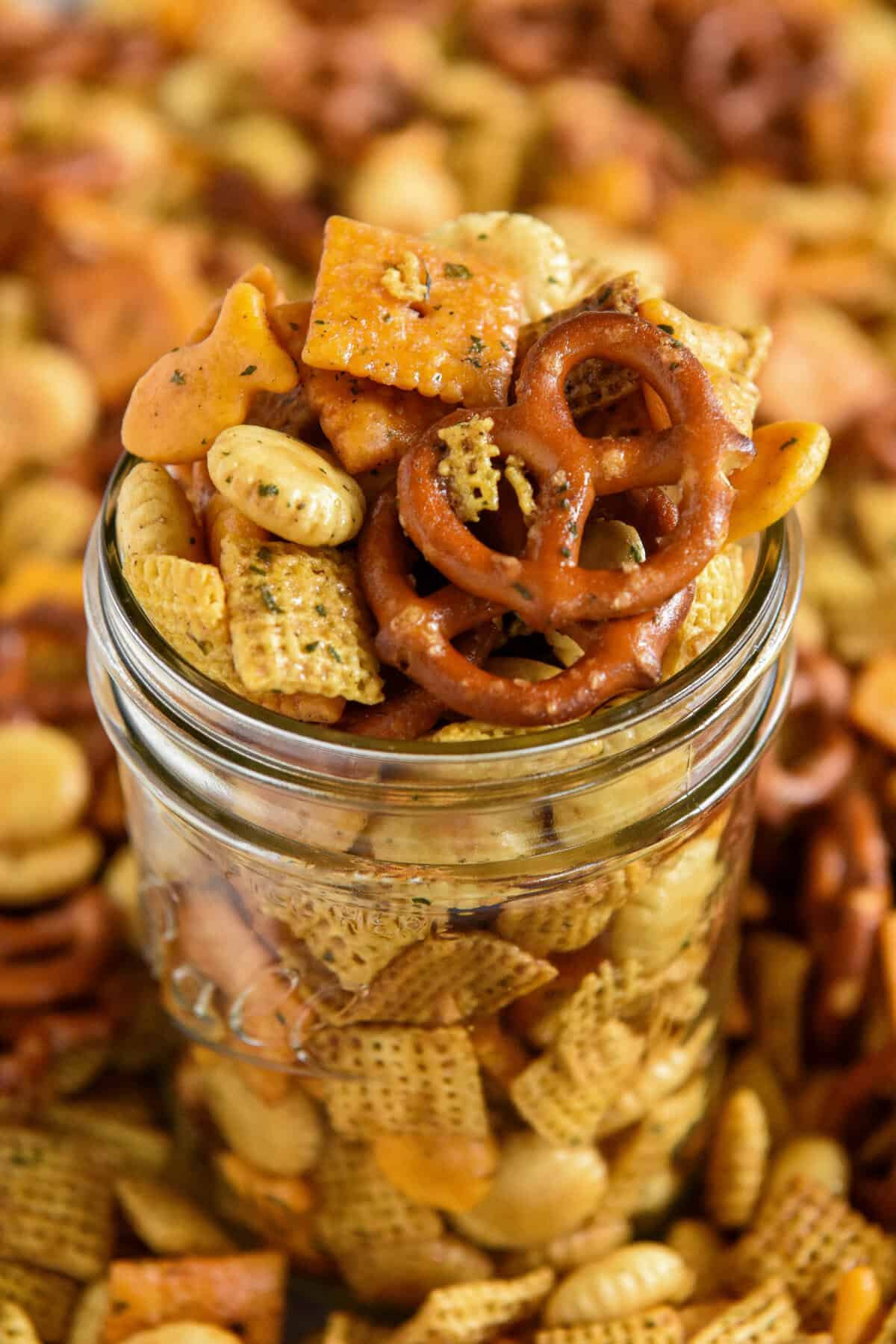 A glass jar filled with cheddar chex mix, including pretzels, cheese crackers, peanuts, and seasoned cereal pieces, with more snack mix scattered in the background.