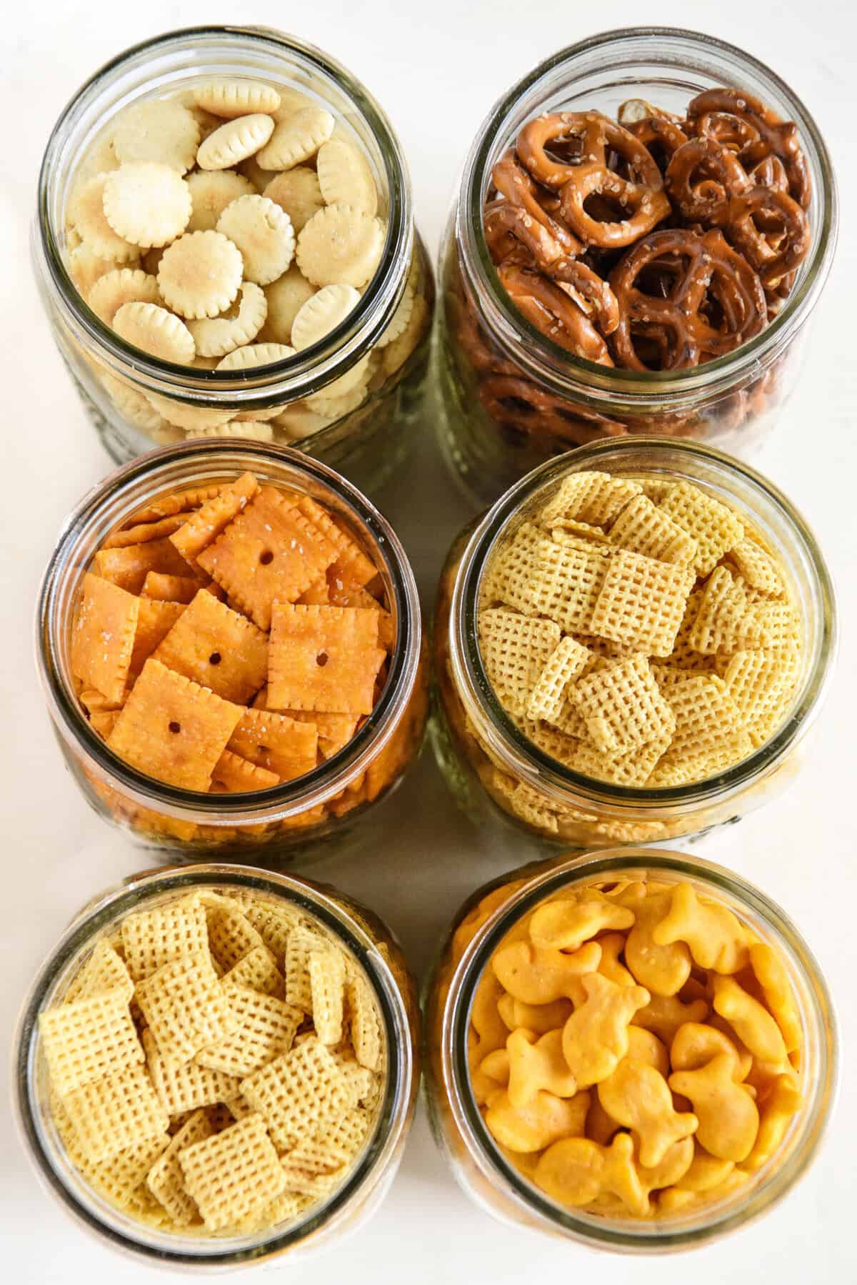 Six glass jars filled with different snacks: mini round crackers, pretzels, square cheese crackers, fish-shaped crackers, and two types of Chex cereal, all arranged neatly on a white surface.