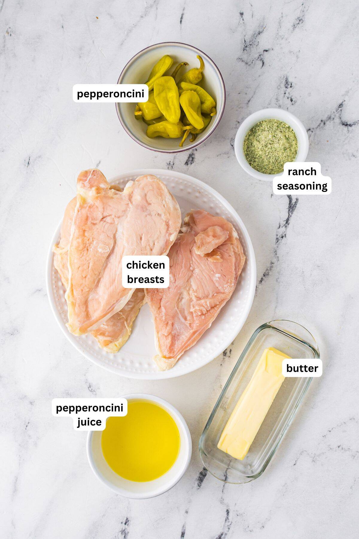 Labeled ingredients on a marble surface: raw chicken breasts on a plate, a bowl of pepperoncini, ranch seasoning in a small bowl, a bowl of pepperoncini juice, and a stick of butter in a butter dish.