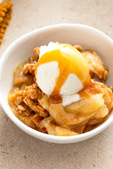A bowl of apple crisp topped with a scoop of vanilla ice cream and drizzled with caramel sauce, served on a beige surface.